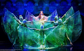 Five performers on stage wear elaborate, shimmery costumes with fan-like extensions, creating a dramatic, symmetrical display against an ornate backdrop with blue and green lighting.