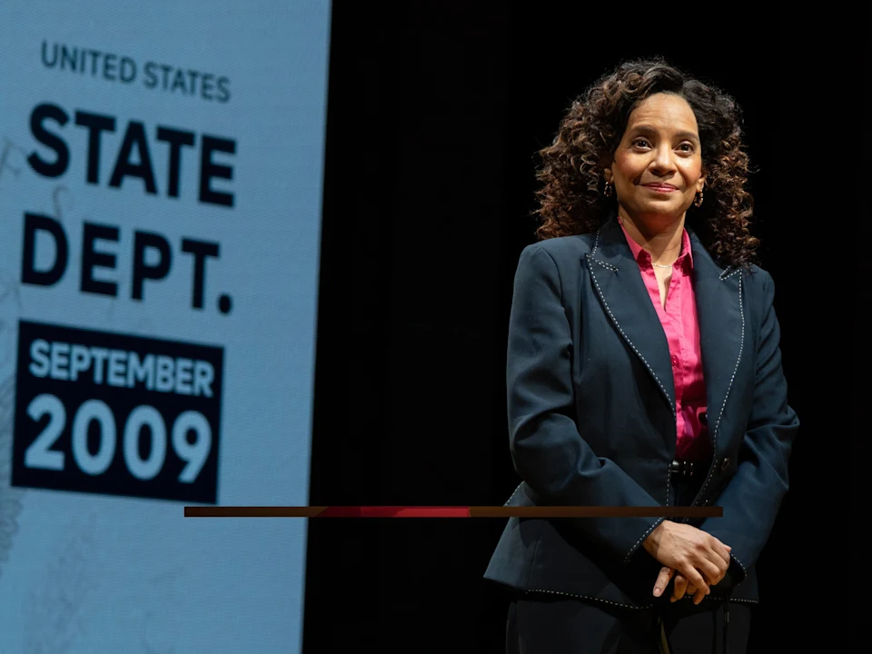 A woman in a suit stands on stage next to a screen displaying "UNITED STATES STATE DEPT. SEPTEMBER 2009.