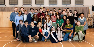 A large group of people of various ages pose together indoors on a gym or rehearsal room floor, smiling at the camera.