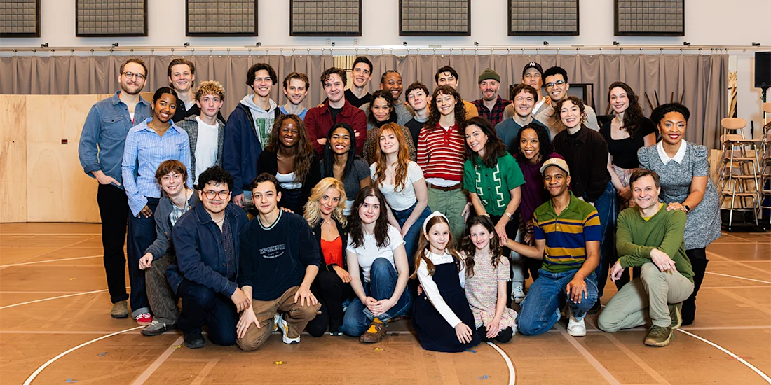 A large group of people of various ages pose together indoors on a gym or rehearsal room floor, smiling at the camera.