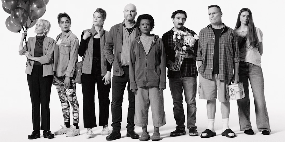 Eight people stand in a row against a white background, each with a neutral expression and casual clothing. One holds balloons, another holds flowers, and another holds a grocery bag.