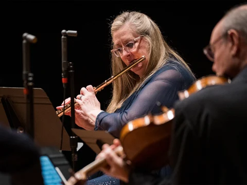 A woman with long blonde hair and glasses plays a flute while reading sheet music. Other musicians with string instruments are also partially visible.