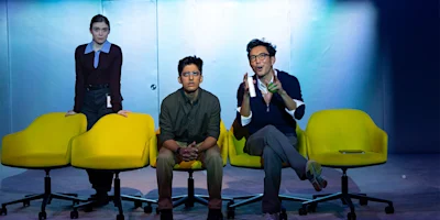 Three people sit and stand by yellow chairs under bright lighting; one person stands behind, another sits in the center, and the third gestures while speaking.