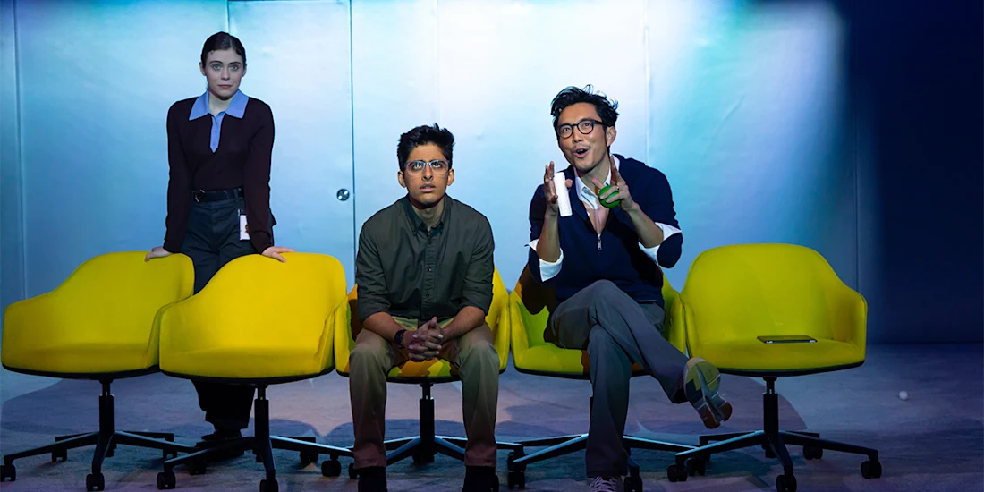 Three people sit and stand by yellow chairs under bright lighting; one person stands behind, another sits in the center, and the third gestures while speaking.