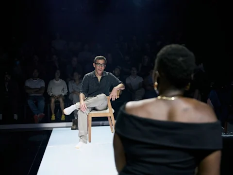 A man sits on a chair facing a woman in an off-the-shoulder top, with an audience watching them in a dimly lit room.