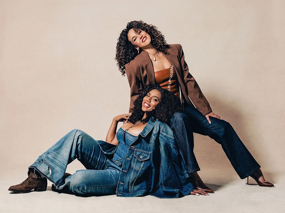 Two women with curly hair pose together; one sits on the floor in denim jeans and jacket, while the other sits behind her in a brown blazer, brown top, and jeans.