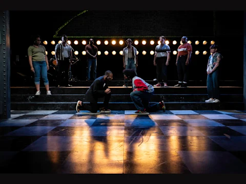 Two people kneel facing each other center stage, while others stand in the background under bright lights on a checkered floor in a theater setting.