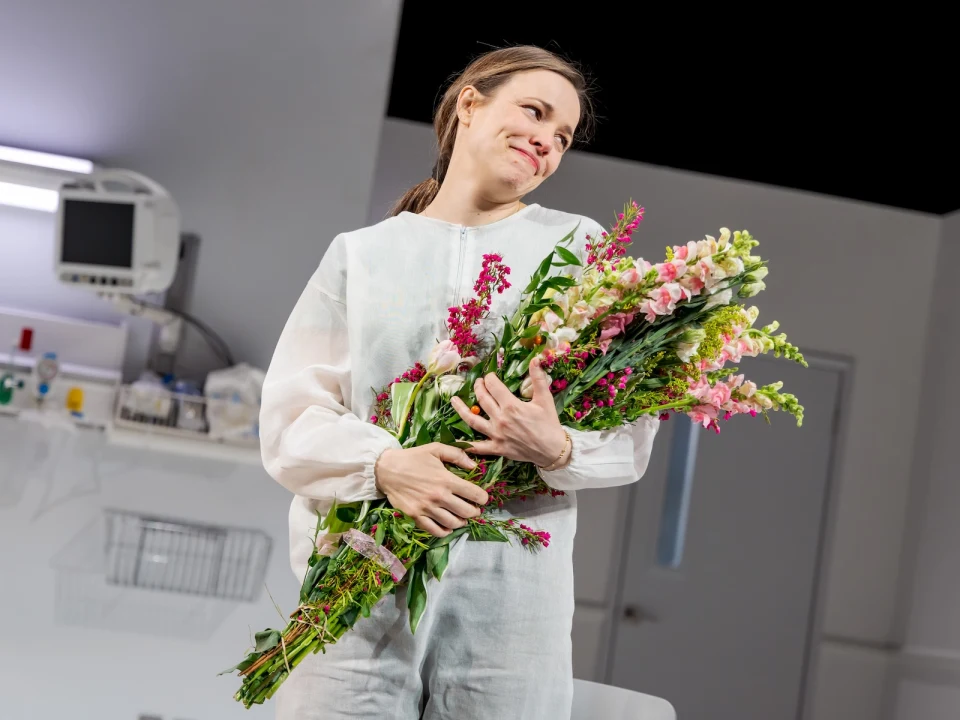 Person in a white outfit holds a large bouquet of flowers in what appears to be a medical or clinical setting.