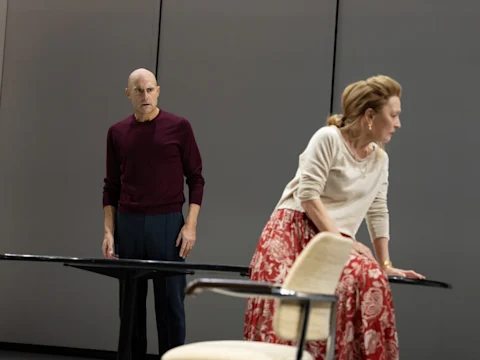 A man stands behind a table looking toward a woman who is leaning on the table and looking away in a minimalist room.