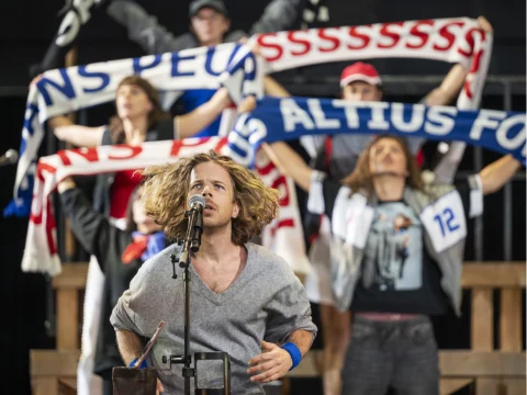 Person with long hair sings into a microphone while a group of people in the background hold up scarves and cheer enthusiastically.