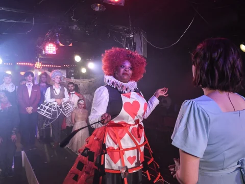 A stage scene with actors in colorful costumes: a person dressed as the Queen of Hearts interacts with another person in blue as a cast stands in the background under theatrical lighting.