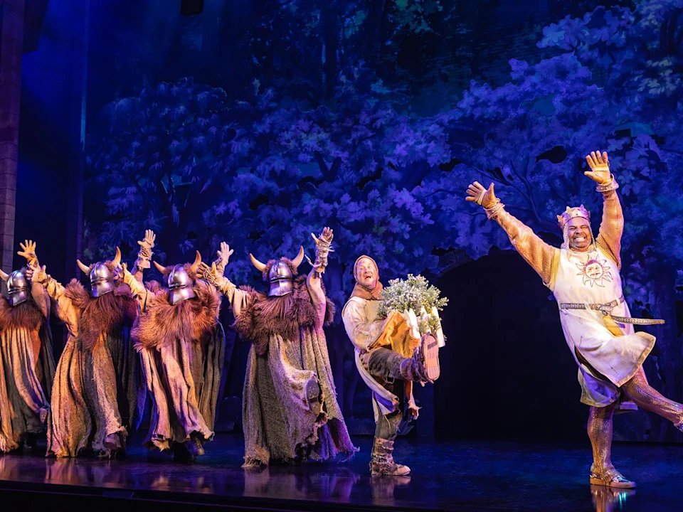 A group of actors in medieval costumes and Viking helmets perform energetically on a stage with a forest backdrop.