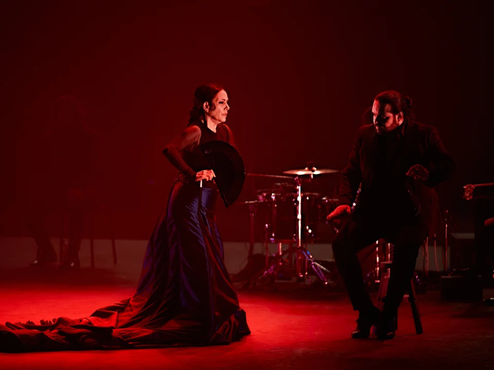 A woman in a long, dark dress holds a black fan, standing and looking at a man who is seated and poised to dance on a stage illuminated by red lighting. Musical instruments are visible in the background.