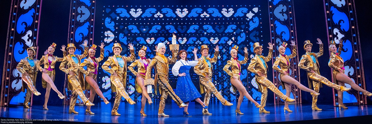 A group of performers in coordinated gold costumes and hats pose in a line on stage with a woman in blue and white at the center, under bright theatrical lighting and ornate set design.