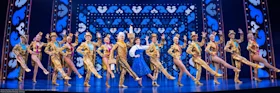 A group of performers in coordinated gold costumes and hats pose in a line on stage with a woman in blue and white at the center, under bright theatrical lighting and ornate set design.