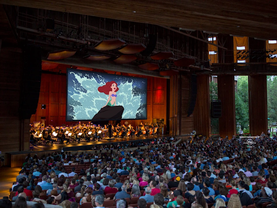 A large audience watches an orchestra perform beneath a big screen displaying a scene from "The Little Mermaid" in a spacious indoor venue.
