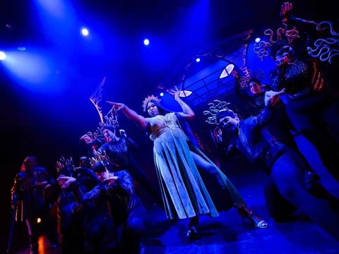 A central performer in a dress gestures dramatically on stage, surrounded by dancers in dark costumes and headpieces under vibrant blue and purple lighting.