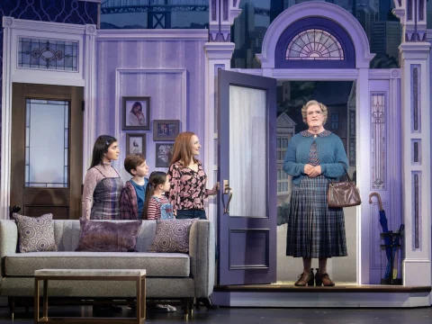 A scene from a stage play shows a family of four inside a living room looking at a woman dressed as an older lady standing in the doorway. The set has a couch and purple-themed decor.