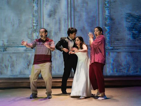 Four people on stage, three in casual clothes and one in a white dress, stand together with surprised expressions against a textured wall backdrop.