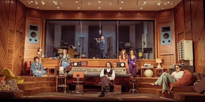 Six people pose in a retro recording studio with wood paneling, large speakers, and a mixing console; one person stands in the sound booth behind glass.