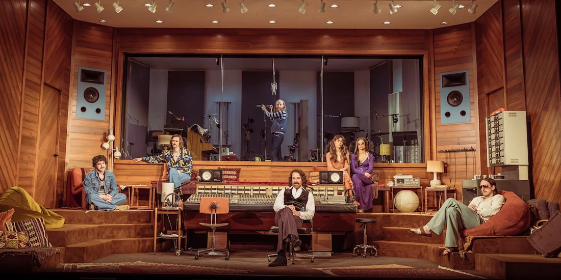 Six people pose in a retro recording studio with wood paneling, large speakers, and a mixing console; one person stands in the sound booth behind glass.