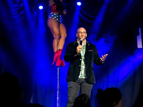 A man holding a microphone stands on stage in front of an audience, while a pole dancer in red boots performs in the background under blue stage lights.