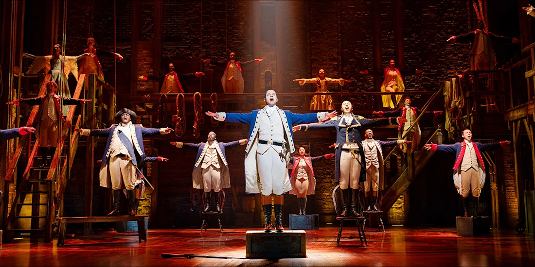 Cast members in historical costumes perform a musical number on stage, with the central figure in blue and others extending their arms outward.