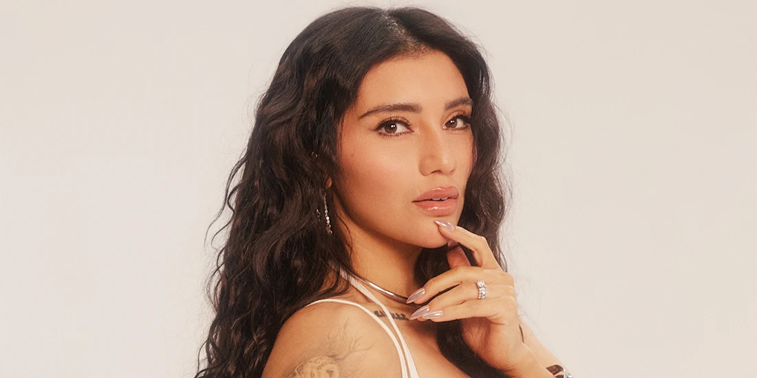 A woman with long wavy dark hair poses against a neutral background, resting her hand near her face and wearing light-colored clothing and minimal jewelry.