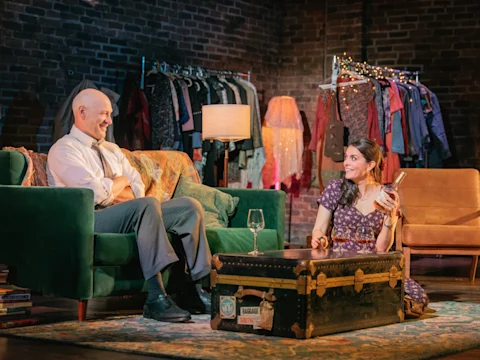 A man and woman sit in a living room set, talking and smiling. The woman holds a liquor bottle and glass. There is a green couch, trunk coffee table, and clothing rack in the background.