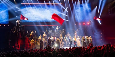 Stage performance with a large cast in period costumes, two people wave red flags, bright stage lights illuminate the scene, and an audience watches in the foreground.