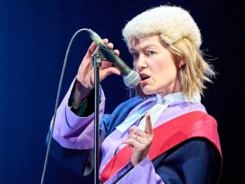 A person wearing a judge's wig and robes speaks into a microphone on stage, holding up one finger while making a point.