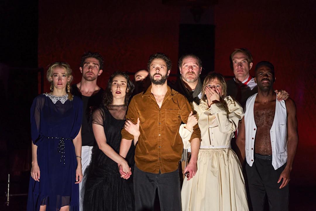 Eight actors stand close together on stage, looking forward with serious and concerned expressions, dressed in a variety of vintage and dramatic costumes.