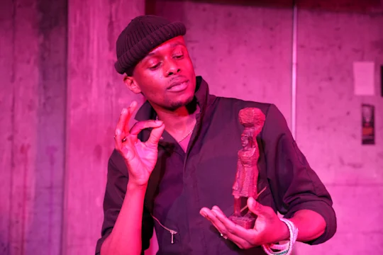 A person wearing a black beanie and jacket holds a small statue in one hand and gestures with the other, standing against a pink-lit concrete wall.