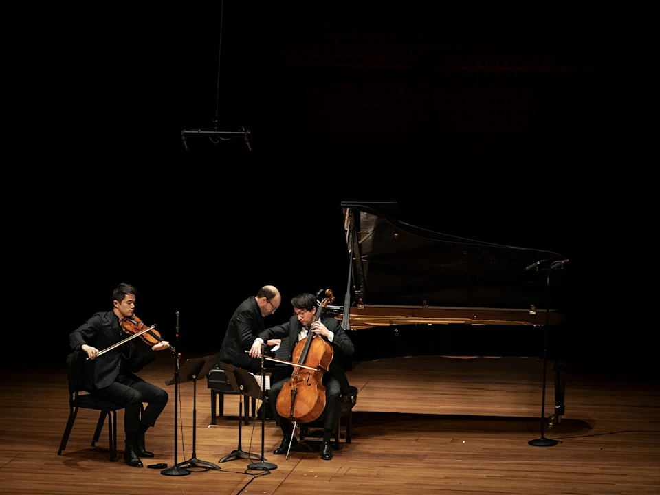 Three musicians in formal wear perform on stage with a violin, cello, and grand piano under spotlight, with microphones set up in front of them.