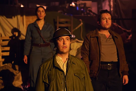 Three people in period clothing stand in a dimly lit room with sandbags and wooden crates, resembling a wartime setting. The person in front wears a military helmet and green jacket.
