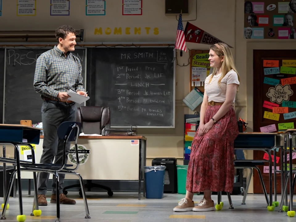 A man stands and talks to a seated young woman in a classroom with desks, a chalkboard, and colorful decorations on the walls.