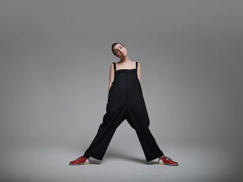 Person stands in a wide stance, wearing an oversized black outfit with red and white shoes against a gray studio backdrop.