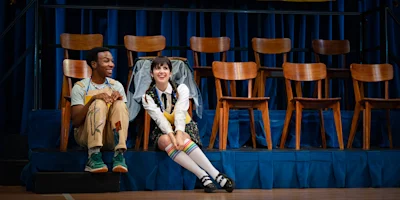 Two people sit onstage in front of a row of empty chairs, smiling and talking. One wears overalls; the other wears a white shirt, vest, and knee-high socks with rainbow stripes.