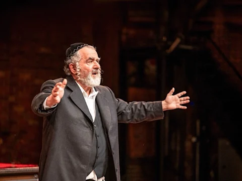 An older man with a beard, wearing a suit and a kippah, stands with arms outstretched, appearing to speak or perform on a dimly lit stage.