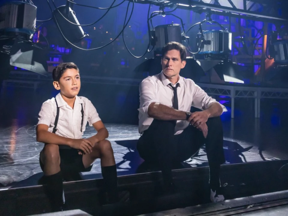 A young boy and an adult man dressed in formal attire sit on the floor of a stage, surrounded by stage lighting equipment.