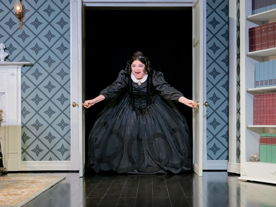 A woman in a long black dress stands in a doorway, leaning forward with arms outstretched, in a room with patterned wallpaper, bookshelves, and a fireplace.