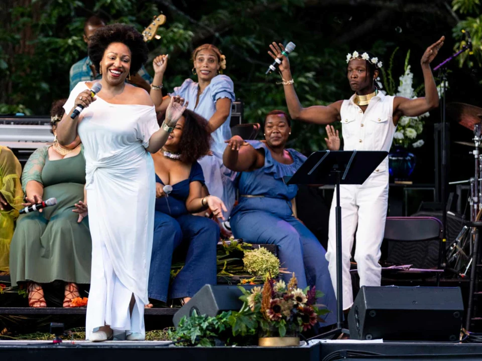 A woman in a white dress sings into a microphone on stage, surrounded by people seated and standing with lively expressions and gestures.