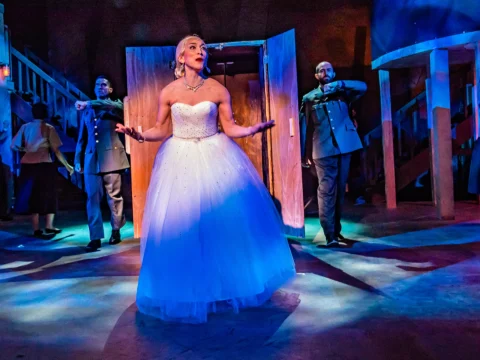 Production shot of Momia en el clóset: Evita's Return in Washington, DC with Fran Tapia as Eva Perón in a white gown surrounded by two men in military uniforms.