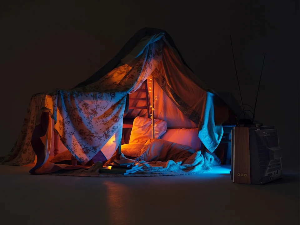 Cozy blanket fort with pillows and a warm glow inside, next to an old television set with antennas, set against a dark background.