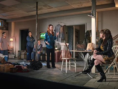 Five women are on stage in a cluttered, domestic set; four stand or sit casually while one woman gestures as if speaking, and another sits apart in a dress and tall boots.