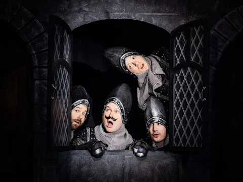 Four people dressed as medieval knights in chainmail and helmets look out of a window with diamond-patterned shutters, making expressive faces.