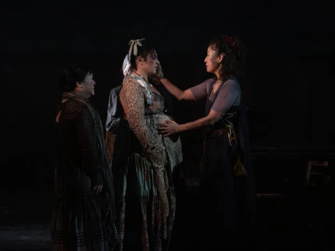 Three individuals in period clothing are on a dimly lit stage, with one person gently touching the face of another, who appears to be pregnant. The third person is observing the interaction.