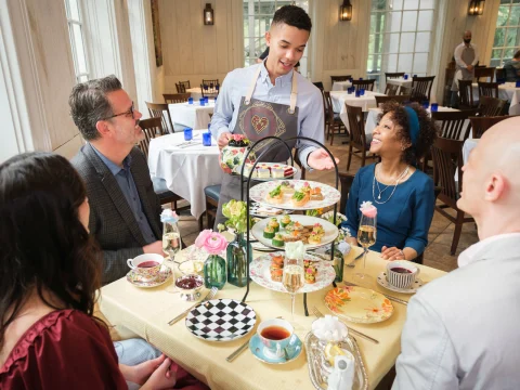 A waiter serves a group of four people seated around a table decorated with a tiered tray of appetizers, flowers, and teacups in a well-lit dining room.