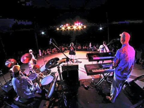 A band performs on a dimly lit stage. The audience is visible in the background. The drummer, keyboardist, and other band members face the crowd under bright stage lights.