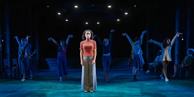 A woman stands center stage under a spotlight while six other actors pose dramatically behind her, all set against a dark, blue-lit theatrical background.